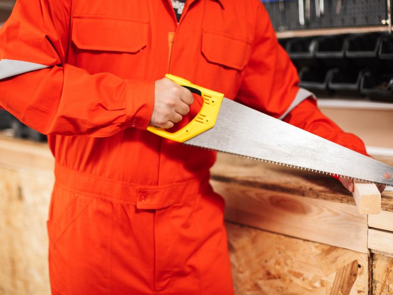 Close up builder in orange work clothes sawing wooden plank with